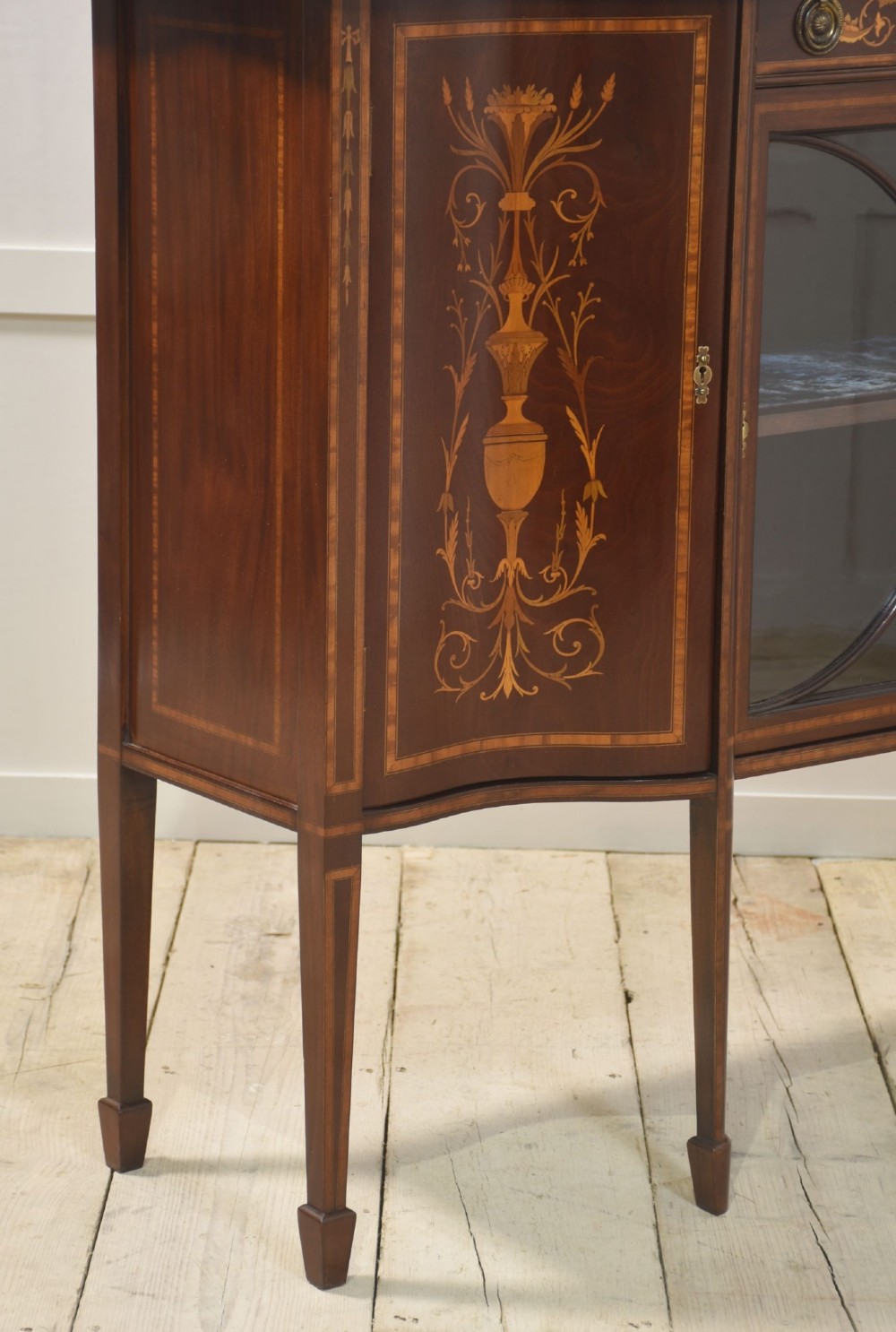 An Edwardian mahogany display cabinet featuring satinwood marquetry inlay, urn motifs, a swan-neck pediment, curved glass doors, and tapered legs. View XII