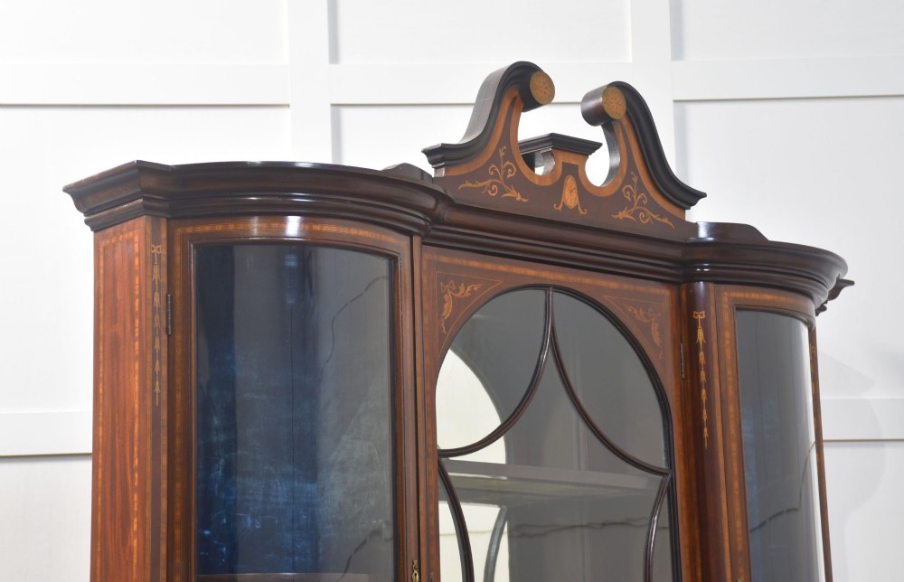 An Edwardian mahogany display cabinet featuring satinwood marquetry inlay, urn motifs, a swan-neck pediment, curved glass doors, and tapered legs. View XIV