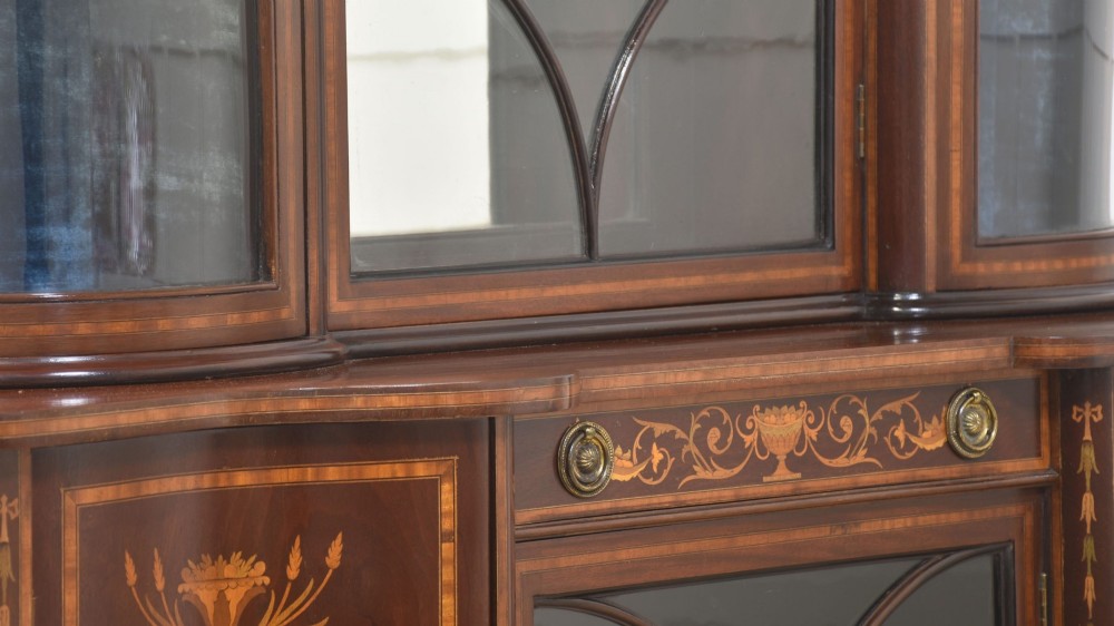 An Edwardian mahogany display cabinet featuring satinwood marquetry inlay, urn motifs, a swan-neck pediment, curved glass doors, and tapered legs. View XV