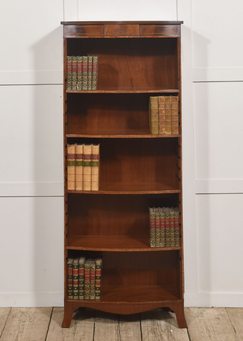 A tall, narrow mahogany open bookcase featuring five shelves, decorative inlay on the top frieze, and shaped bracket feet.