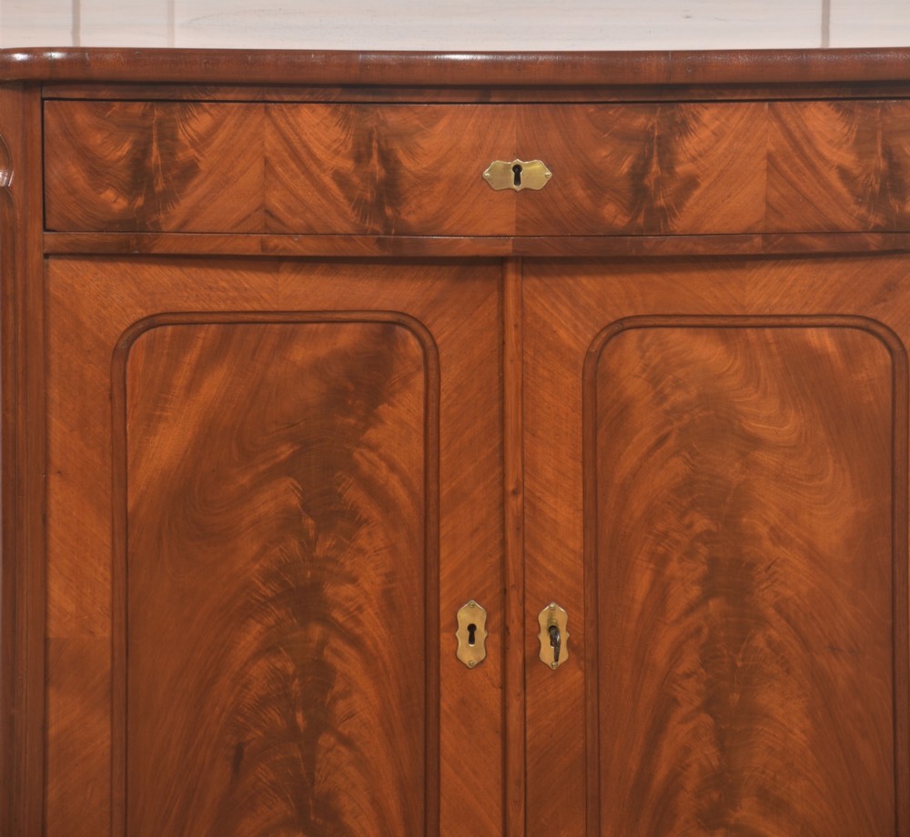 A 19th-century flame mahogany side cabinet with a single top drawer above two panelled cupboard doors with brass keyhole escutcheons. View III