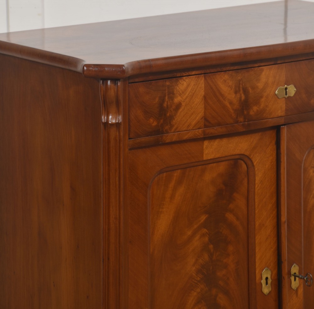 A 19th-century flame mahogany side cabinet with a single top drawer above two panelled cupboard doors with brass keyhole escutcheons. View IV