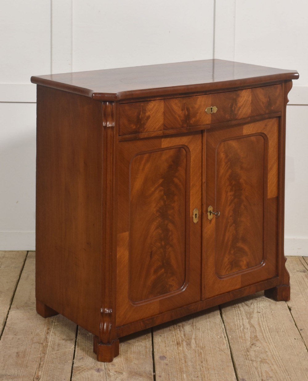 A 19th-century flame mahogany side cabinet with a single top drawer above two panelled cupboard doors with brass keyhole escutcheons. View V