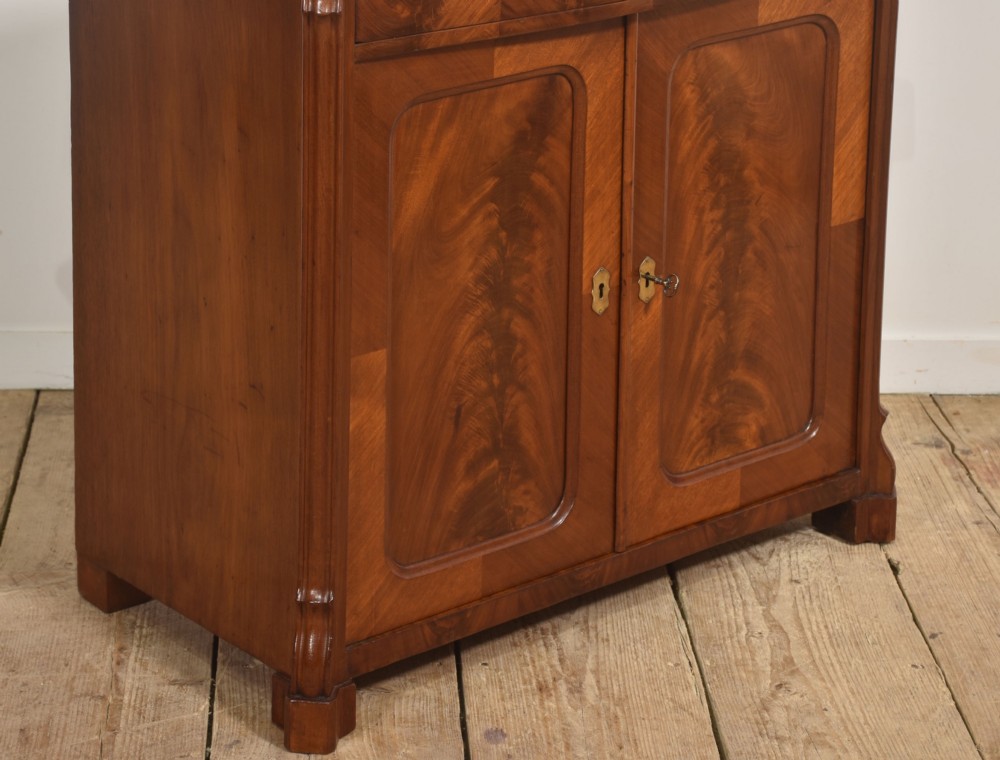 A 19th-century flame mahogany side cabinet with a single top drawer above two panelled cupboard doors with brass keyhole escutcheons. View VII