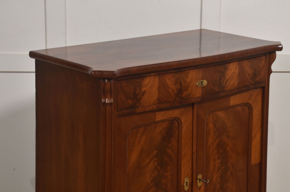A 19th-century flame mahogany side cabinet with a single top drawer above two panelled cupboard doors with brass keyhole escutcheons. View VIII