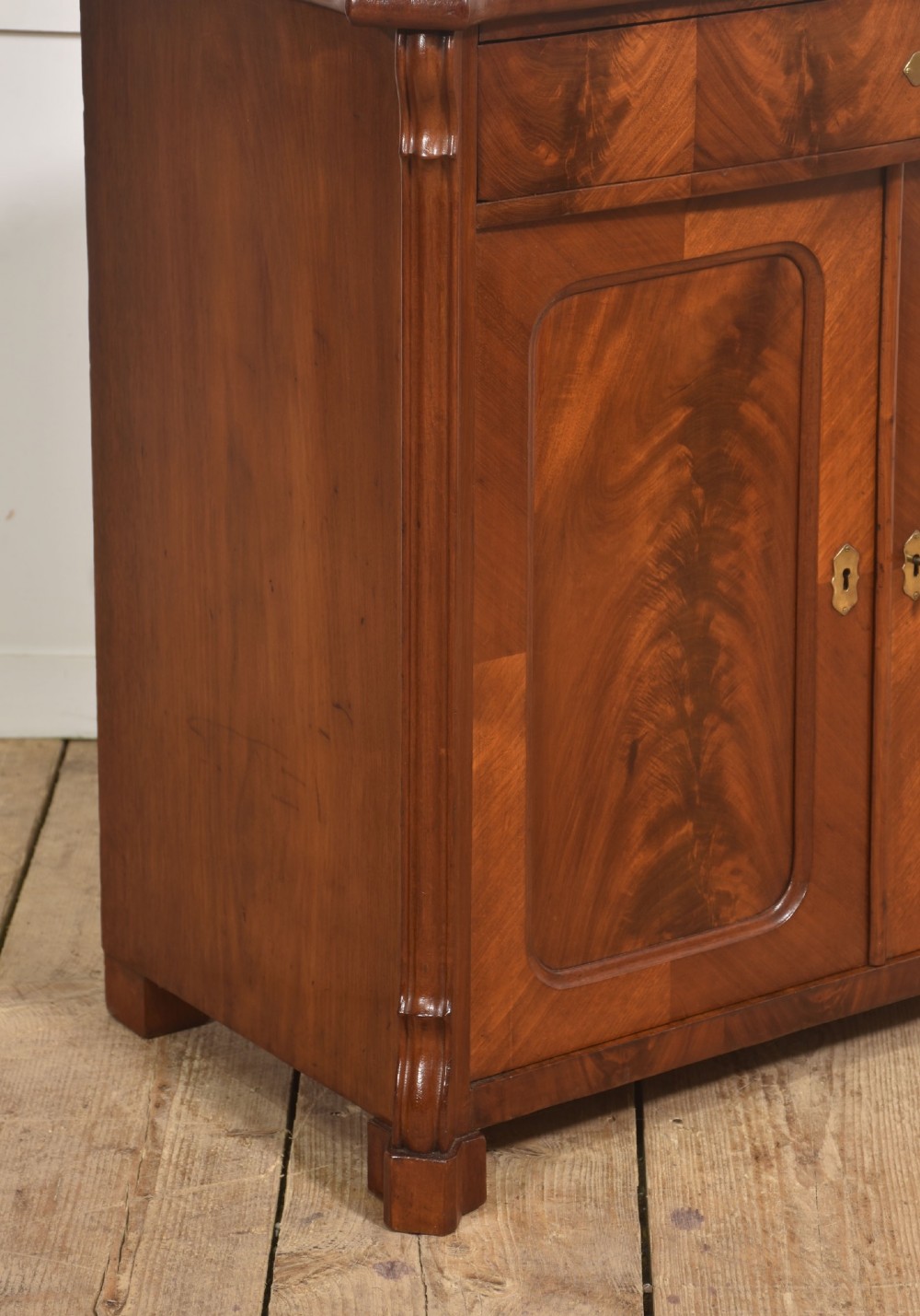 A 19th-century flame mahogany side cabinet with a single top drawer above two panelled cupboard doors with brass keyhole escutcheons. View IX