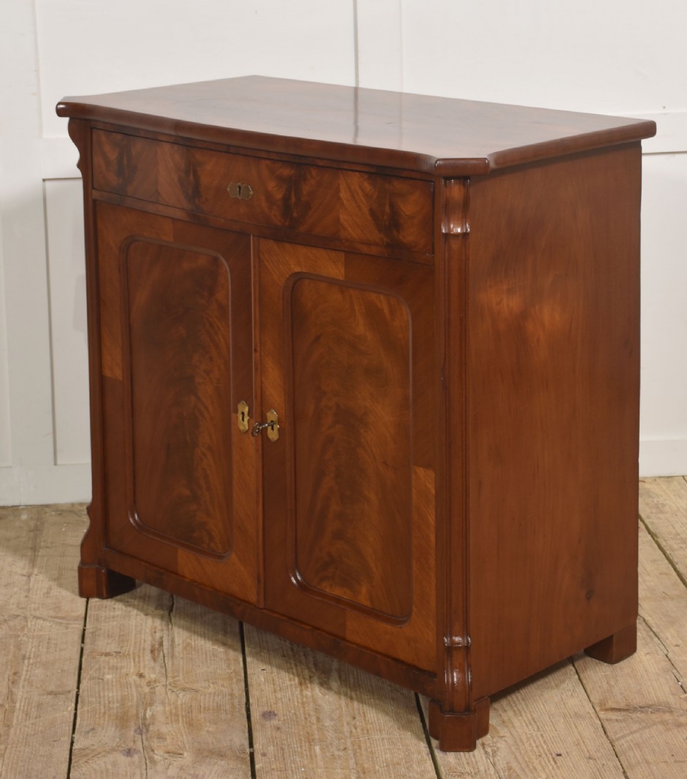 A 19th-century flame mahogany side cabinet with a single top drawer above two panelled cupboard doors with brass keyhole escutcheons. View X