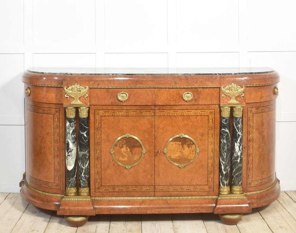 A French bow-front buffet sideboard featuring a dark marble top, burr walnut marquetry, central figural medallions, and flanking marble columns with gilt mounts.