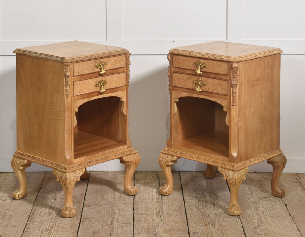 A pair of golden burr walnut bedside cabinets featuring two upper drawers with brass drop handles above an open cubby, set on carved cabriole legs with claw and ball feet.