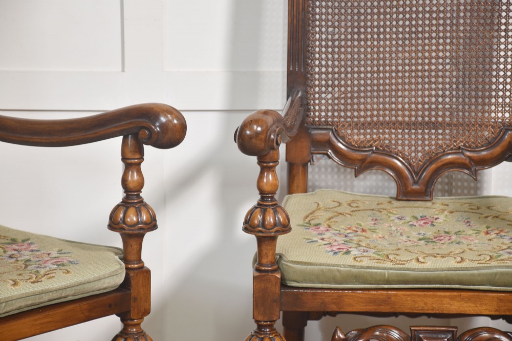 A pair of high-backed carved walnut open armchairs featuring tall cane panels, turned supports, intricate scrolling stretchers, and floral needlepoint cushions. View IV