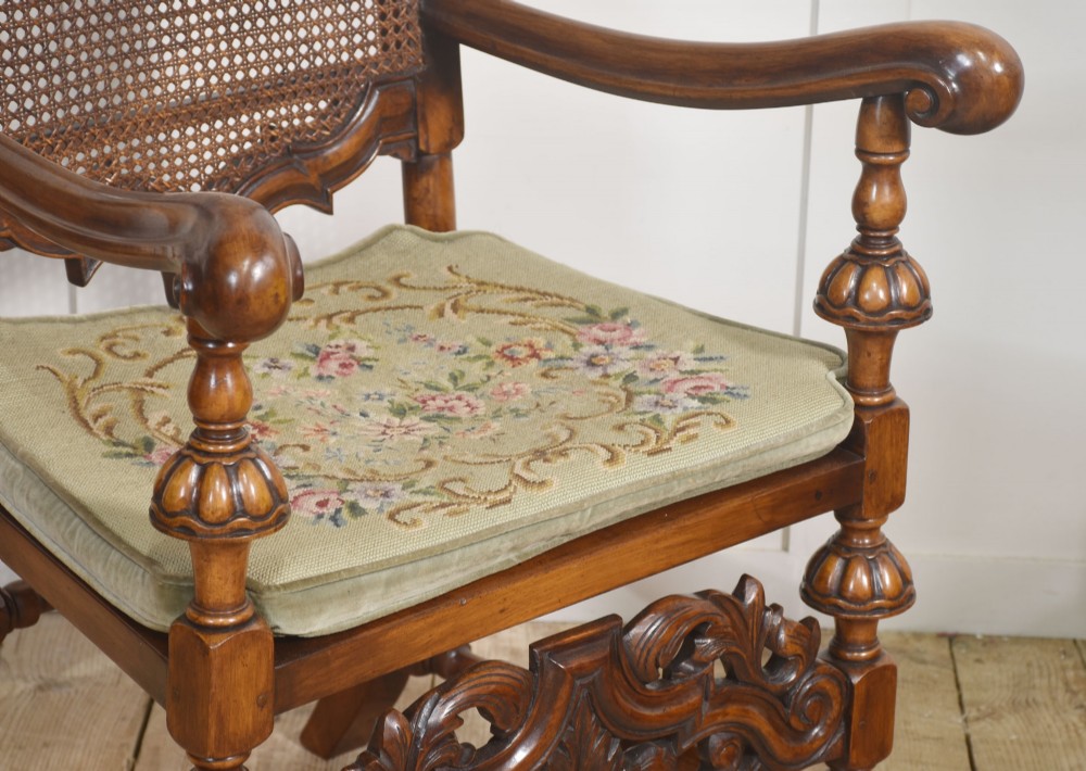 A pair of high-backed carved walnut open armchairs featuring tall cane panels, turned supports, intricate scrolling stretchers, and floral needlepoint cushions. View IX