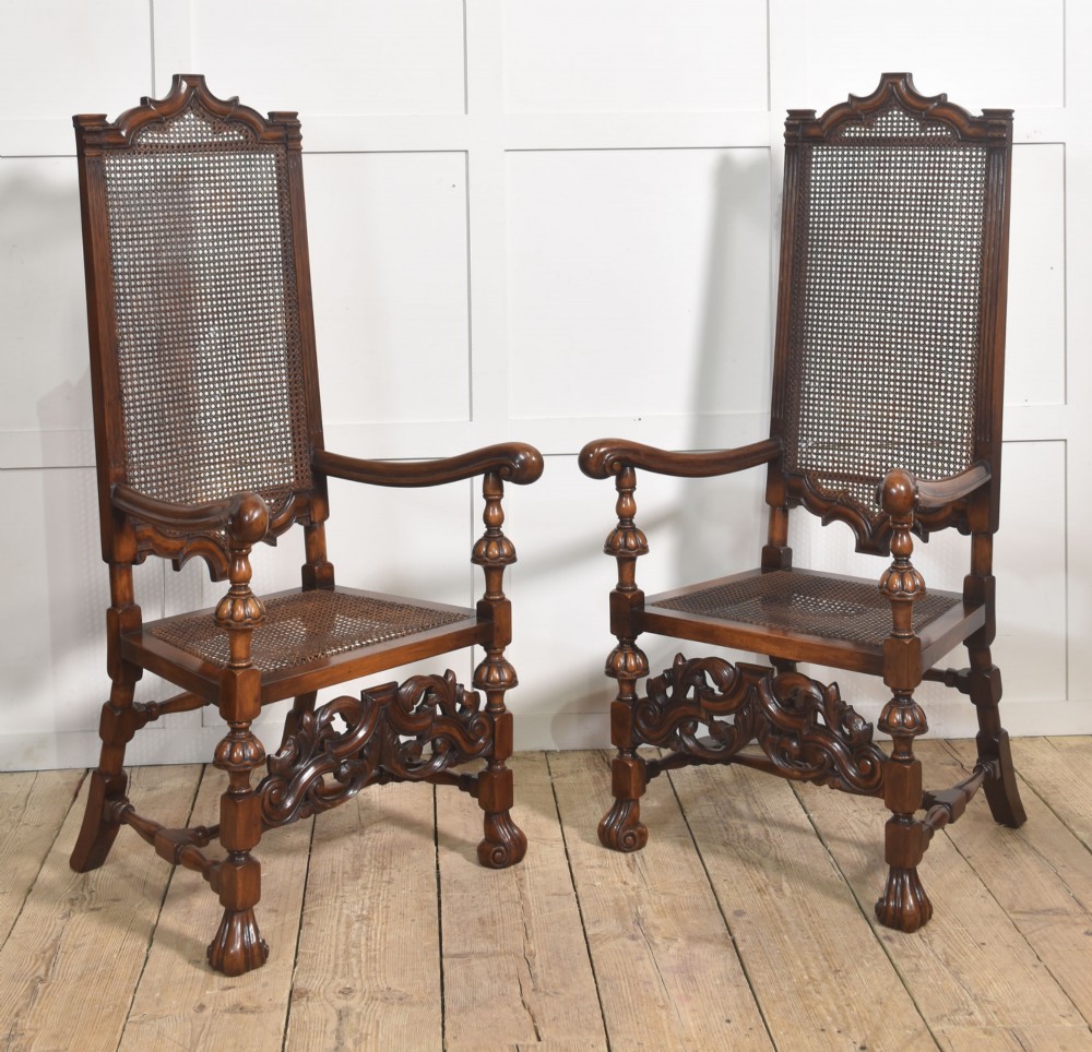 A pair of high-backed carved walnut open armchairs featuring tall cane panels, turned supports, intricate scrolling stretchers, and floral needlepoint cushions. View X