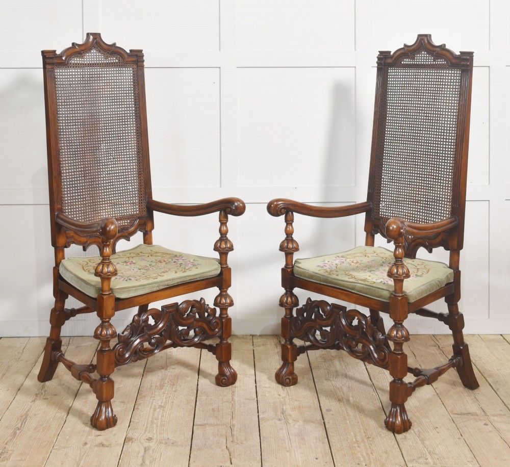 A pair of high-backed carved walnut open armchairs featuring tall cane panels, turned supports, intricate scrolling stretchers, and floral needlepoint cushions.