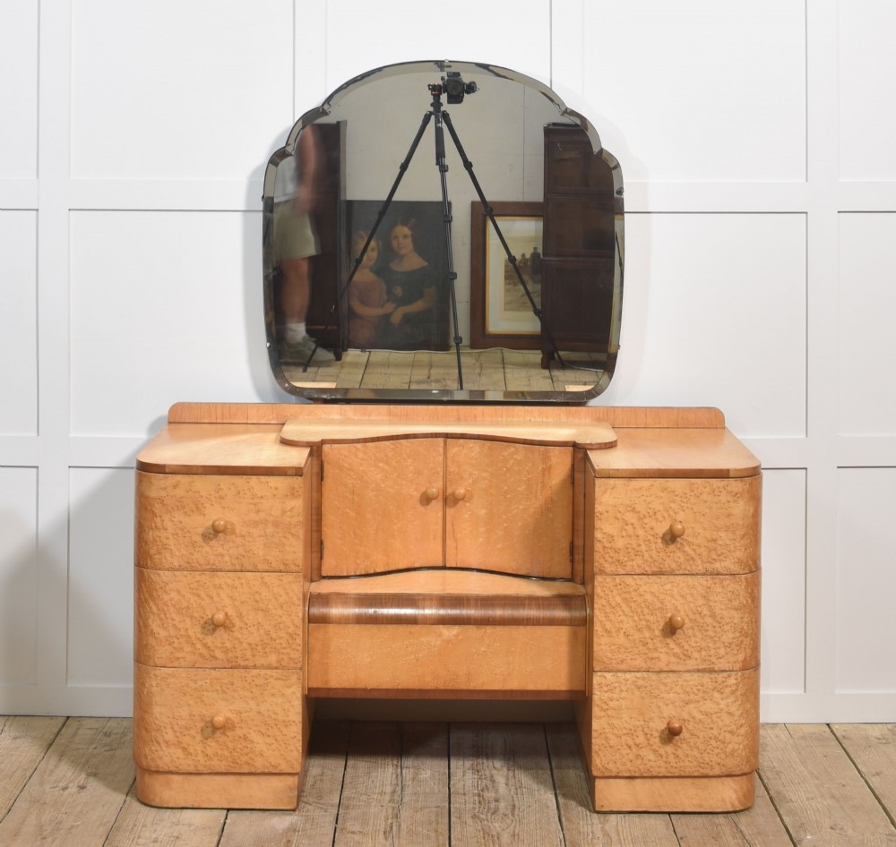 An Art Deco light blonde birds eye maple dressing table featuring curved pedestal drawers, a central cupboard, and a large shaped mirror attached above.