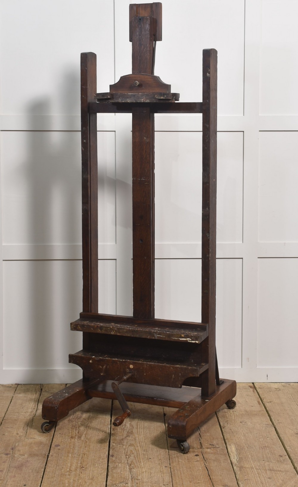 A dark oak adjustable artist studio easel featuring a hand-crank winding mechanism and a castored base.