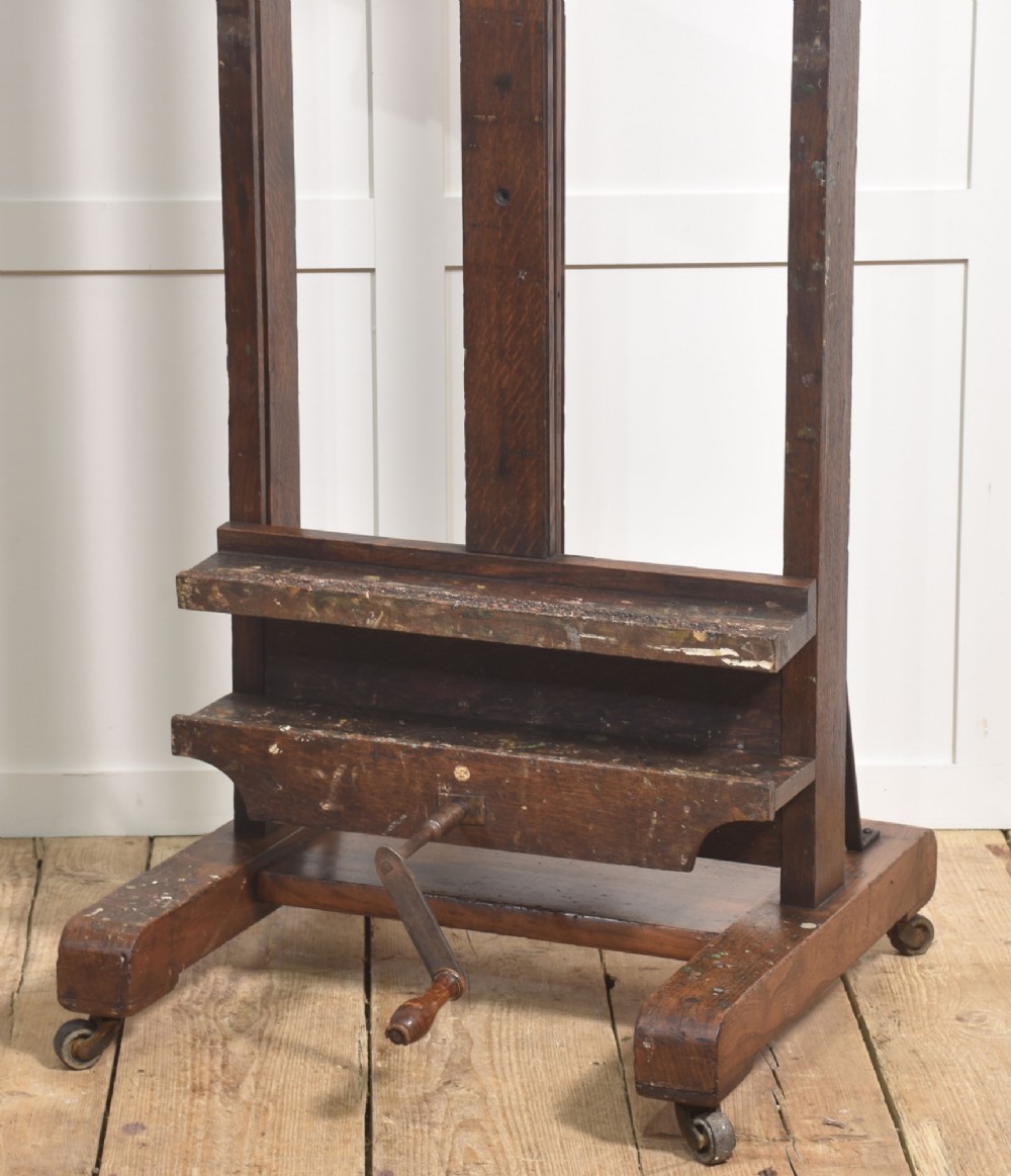 A dark oak adjustable artist studio easel featuring a hand-crank winding mechanism and a castored base. View V