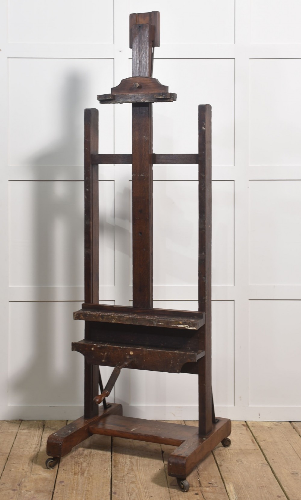 A dark oak adjustable artist studio easel featuring a hand-crank winding mechanism and a castored base. View VI