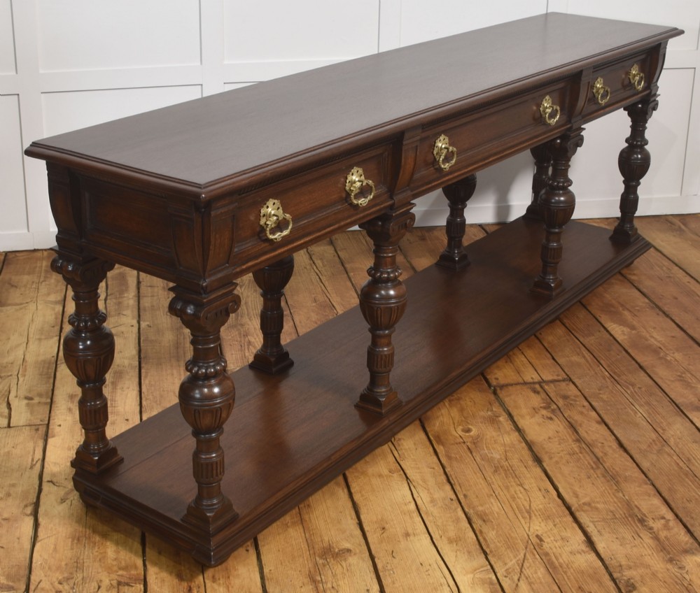 A dark oak Jacobean style dresser base featuring three drawers with decorative brass handles, turned baluster legs, and a full-length lower pot board shelf. View V
