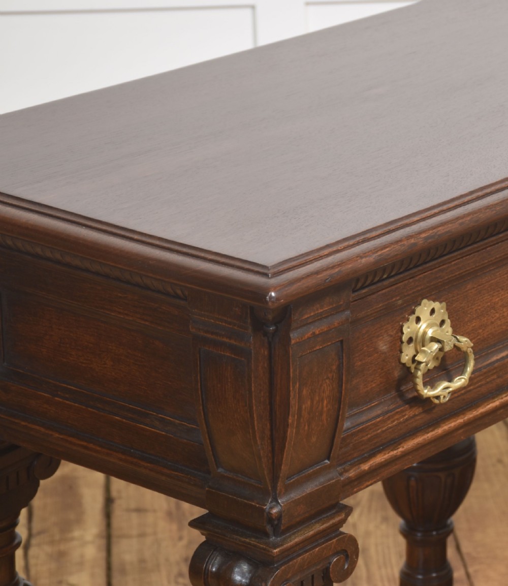 A dark oak Jacobean style dresser base featuring three drawers with decorative brass handles, turned baluster legs, and a full-length lower pot board shelf. View VI