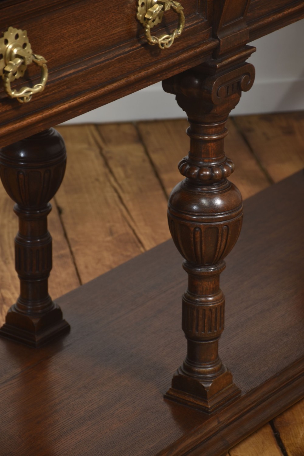 A dark oak Jacobean style dresser base featuring three drawers with decorative brass handles, turned baluster legs, and a full-length lower pot board shelf. View VII