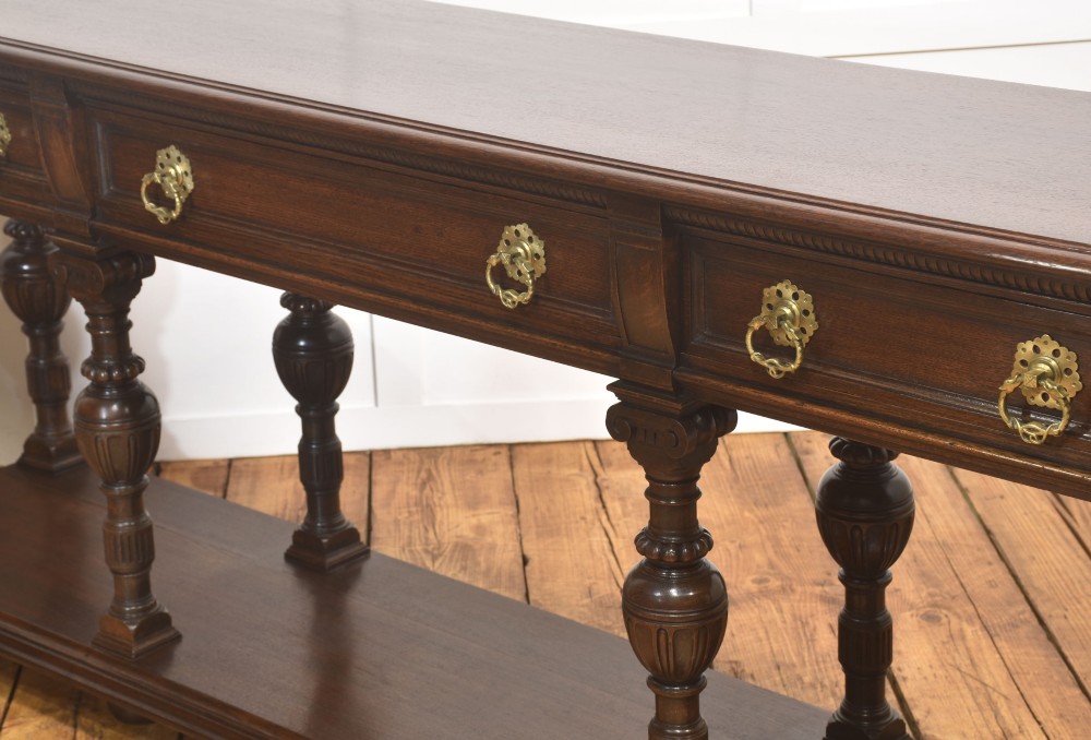 A dark oak Jacobean style dresser base featuring three drawers with decorative brass handles, turned baluster legs, and a full-length lower pot board shelf. View IX