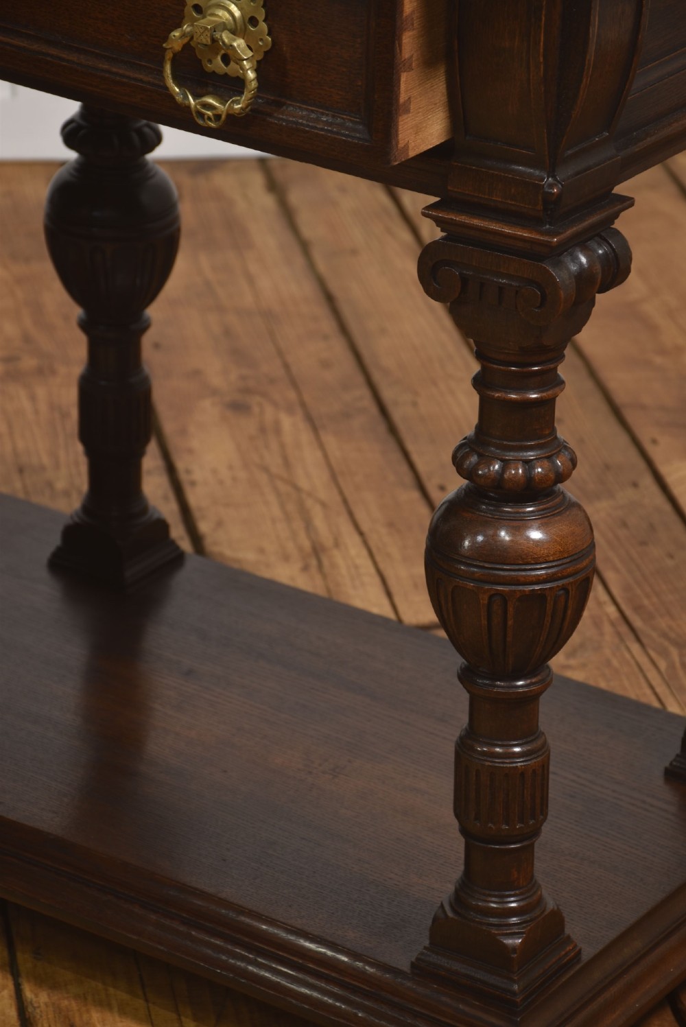 A dark oak Jacobean style dresser base featuring three drawers with decorative brass handles, turned baluster legs, and a full-length lower pot board shelf. View X