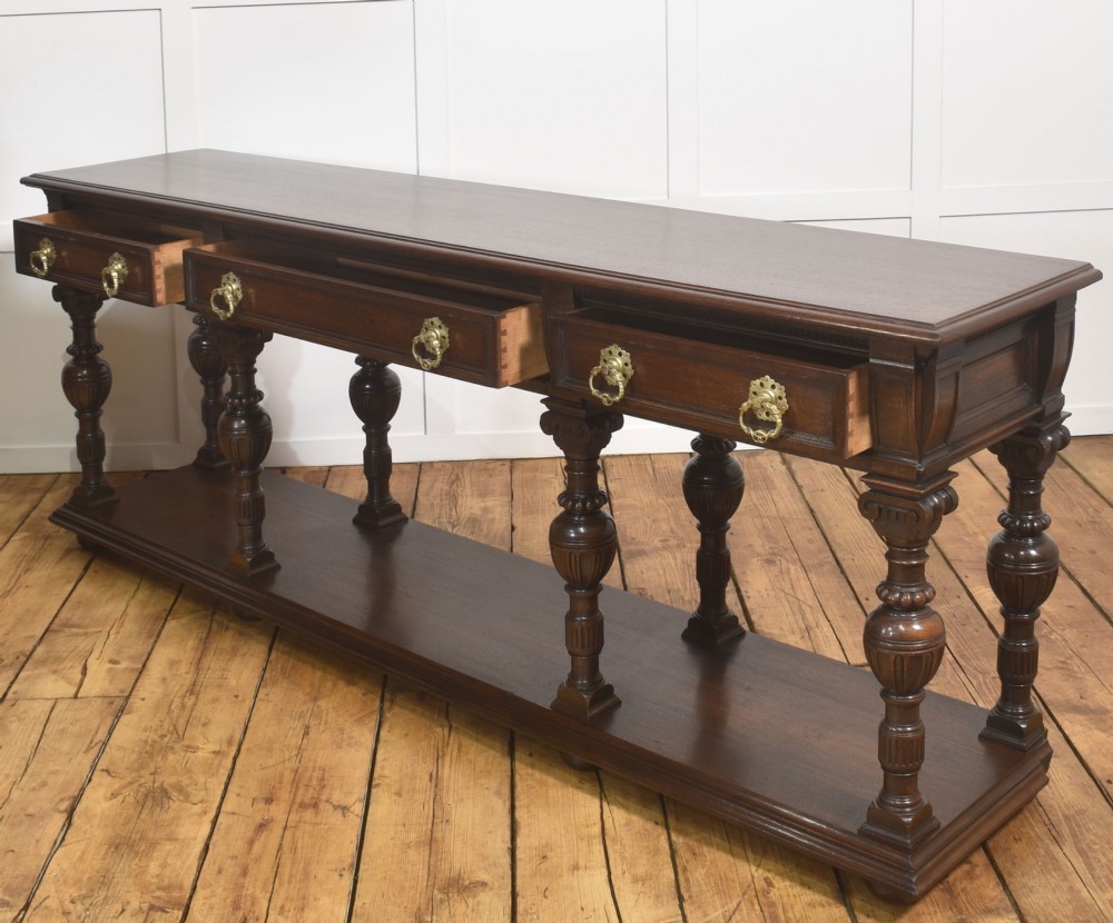 A dark oak Jacobean style dresser base featuring three drawers with decorative brass handles, turned baluster legs, and a full-length lower pot board shelf. View XI