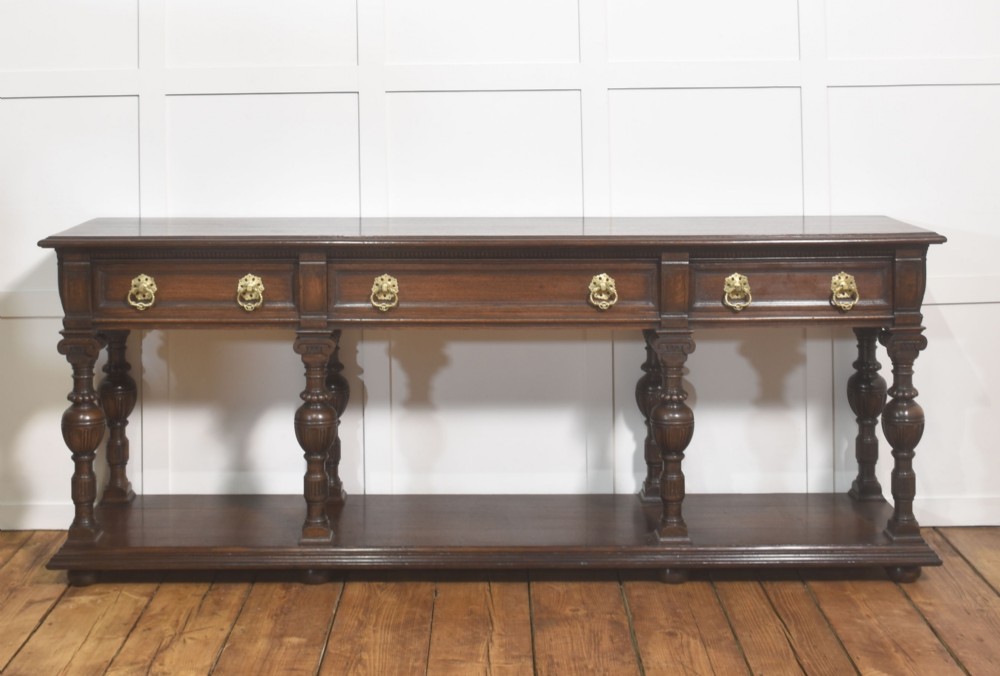 A dark oak Jacobean style dresser base featuring three drawers with decorative brass handles, turned baluster legs, and a full-length lower pot board shelf.