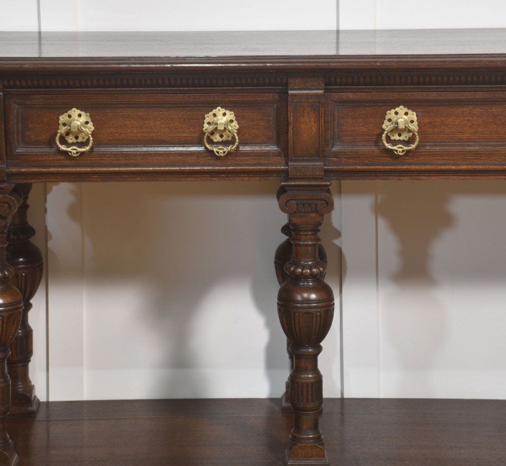 A dark oak Jacobean style dresser base featuring three drawers with decorative brass handles, turned baluster legs, and a full-length lower pot board shelf. View XIV