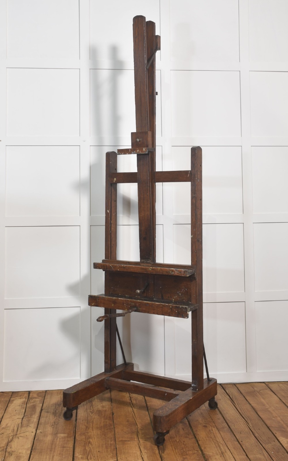A late nineteenth-century dark oak artist studio easel featuring an adjustable central shaft, crank handle, and cast iron castors on an H-frame base. View X