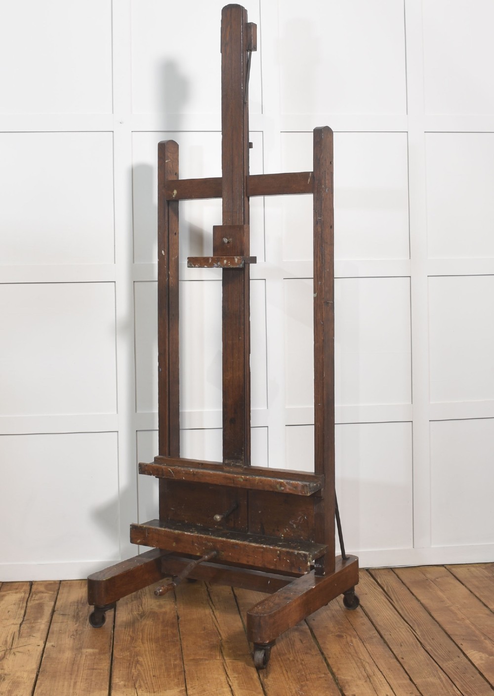 A late nineteenth-century dark oak artist studio easel featuring an adjustable central shaft, crank handle, and cast iron castors on an H-frame base.