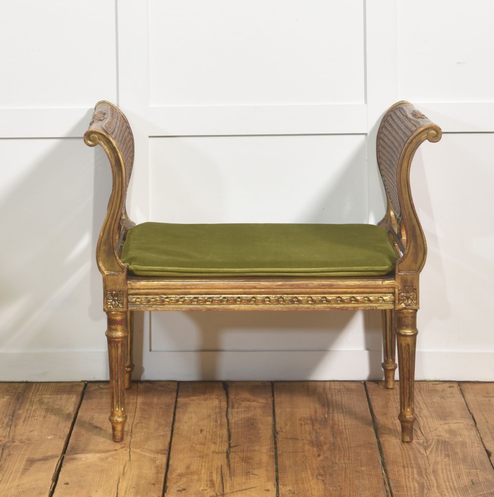 A Louis XVI style giltwood window stool featuring raised caned sides, fluted tapered legs, carved rosette accents, and a fitted olive green velvet cushion.