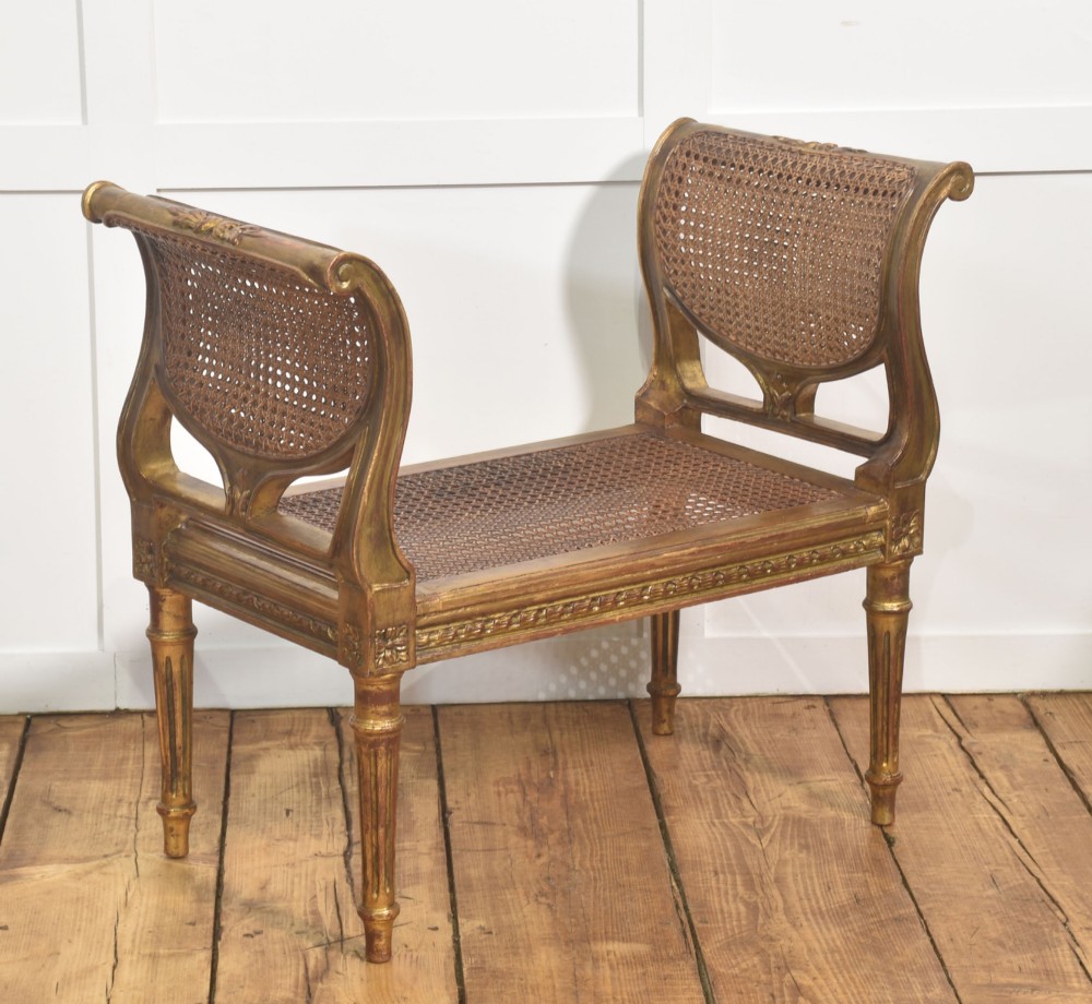 A Louis XVI style giltwood window stool featuring raised caned sides, fluted tapered legs, carved rosette accents, and a fitted olive green velvet cushion. View IV