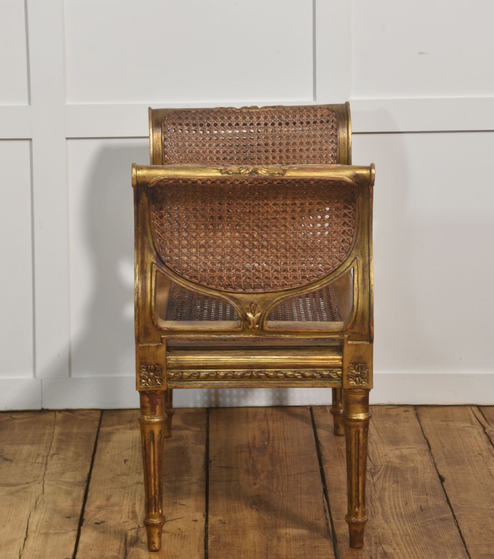 A Louis XVI style giltwood window stool featuring raised caned sides, fluted tapered legs, carved rosette accents, and a fitted olive green velvet cushion. View VI