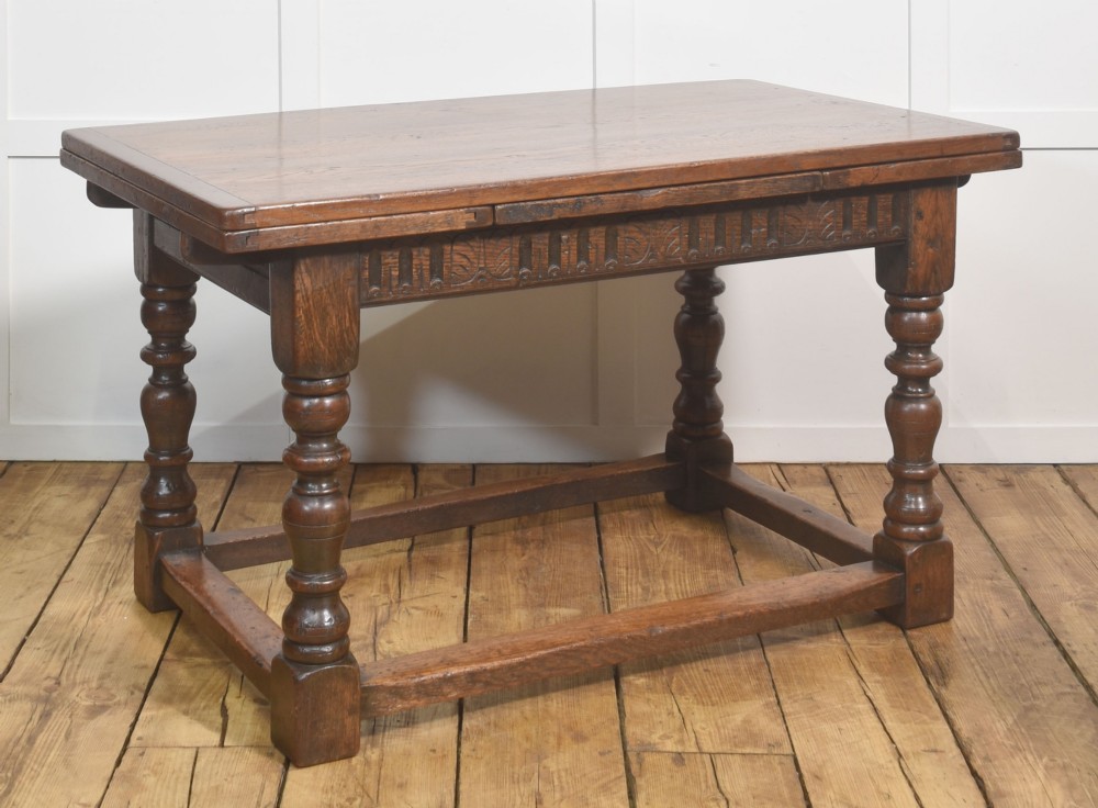 A dark oak Jacobean style draw-leaf dining table featuring carved frieze detailing along the apron, turned baluster legs, and a sturdy low box stretcher base. View III