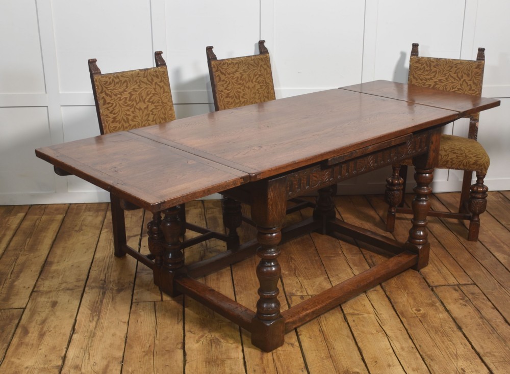 A dark oak Jacobean style draw-leaf dining table featuring carved frieze detailing along the apron, turned baluster legs, and a sturdy low box stretcher base. View X