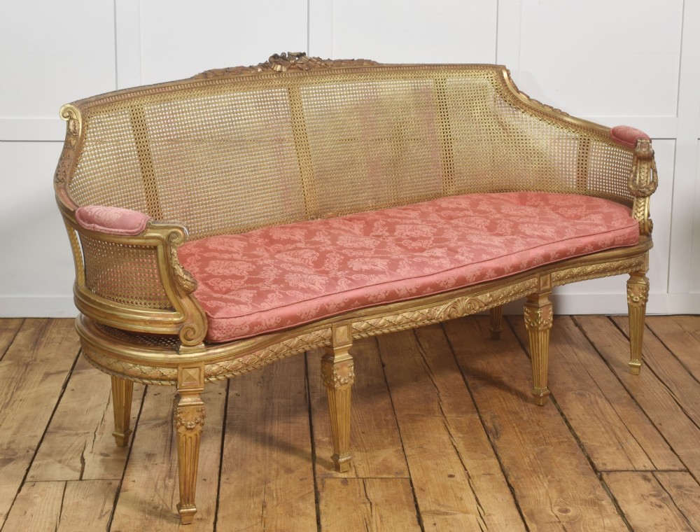 A French Louis XVI style giltwood settee featuring a woven cane backrest, carved fluted legs, and a rose-pink damask upholstered seat cushion. View VI