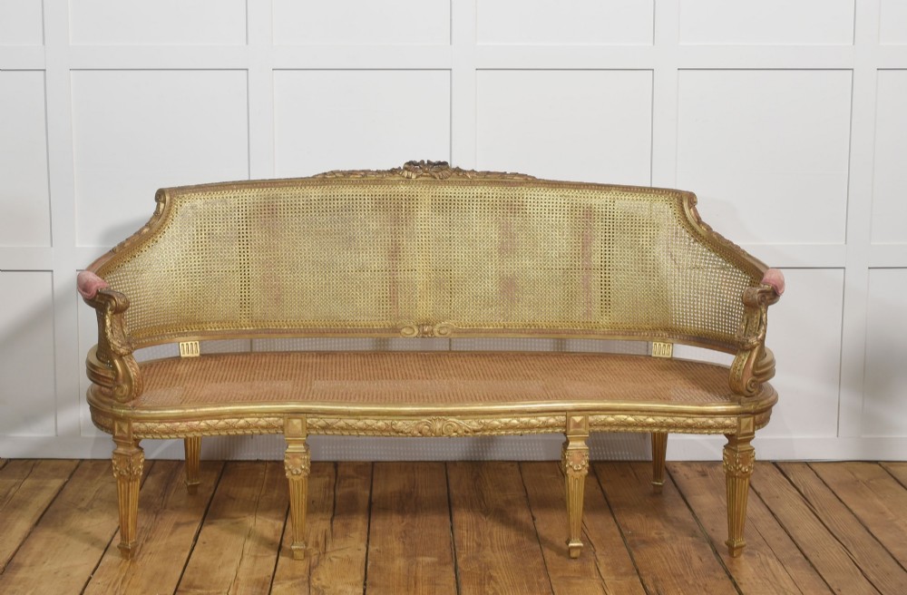 A French Louis XVI style giltwood settee featuring a woven cane backrest, carved fluted legs, and a rose-pink damask upholstered seat cushion. View X