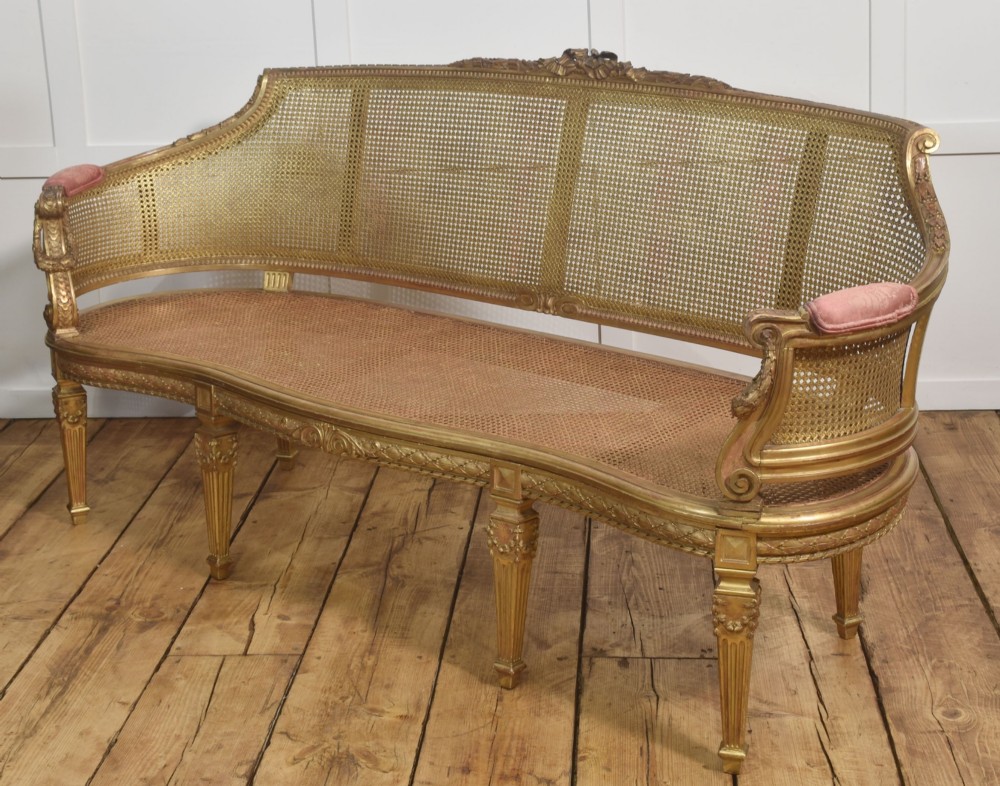 A French Louis XVI style giltwood settee featuring a woven cane backrest, carved fluted legs, and a rose-pink damask upholstered seat cushion. View XII