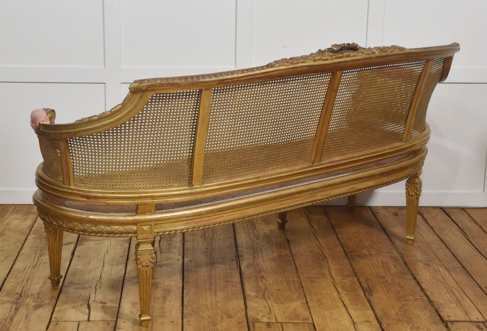 A French Louis XVI style giltwood settee featuring a woven cane backrest, carved fluted legs, and a rose-pink damask upholstered seat cushion. View XIV
