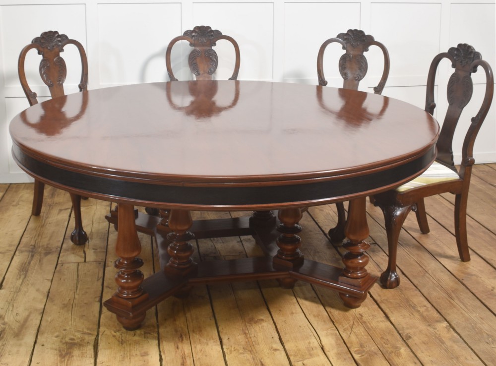 A large circular mahogany centre dining table with a polished surface, dark ebonised apron, and multiple turned legs supported by a cross-stretcher base. View III