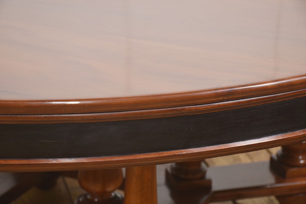A large circular mahogany centre dining table with a polished surface, dark ebonised apron, and multiple turned legs supported by a cross-stretcher base. View V