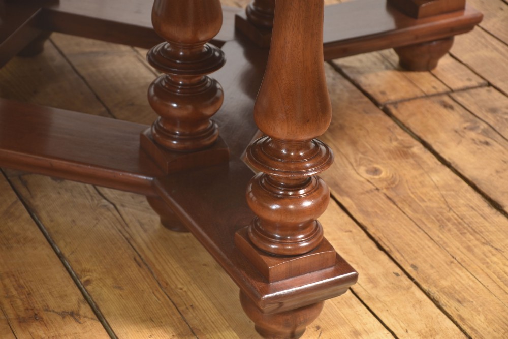 A large circular mahogany centre dining table with a polished surface, dark ebonised apron, and multiple turned legs supported by a cross-stretcher base. View VI