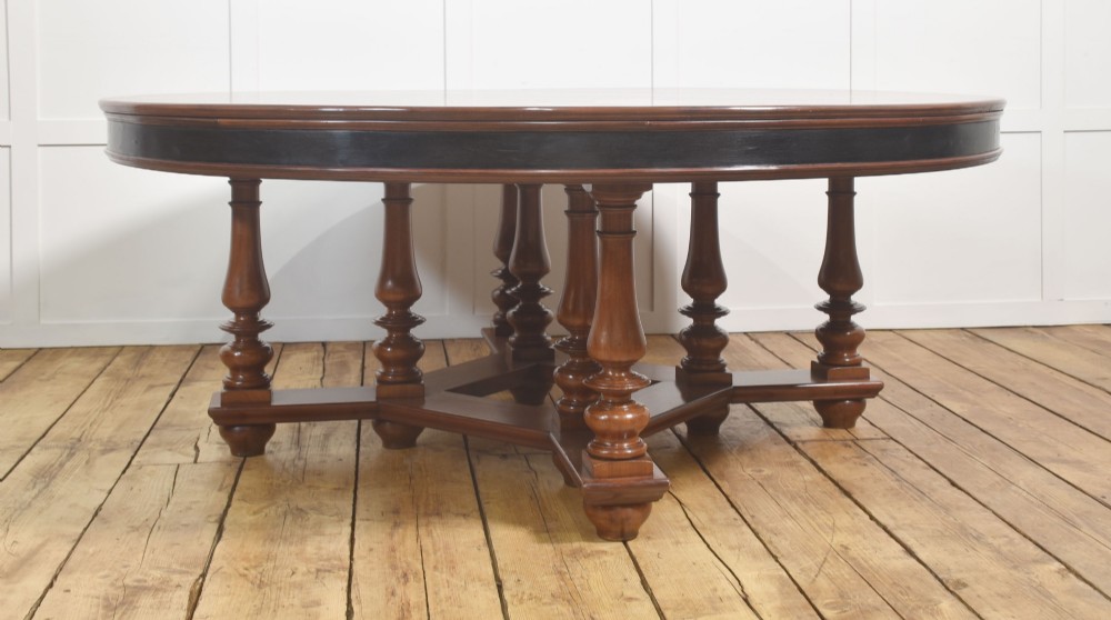 A large circular mahogany centre dining table with a polished surface, dark ebonised apron, and multiple turned legs supported by a cross-stretcher base. View VII