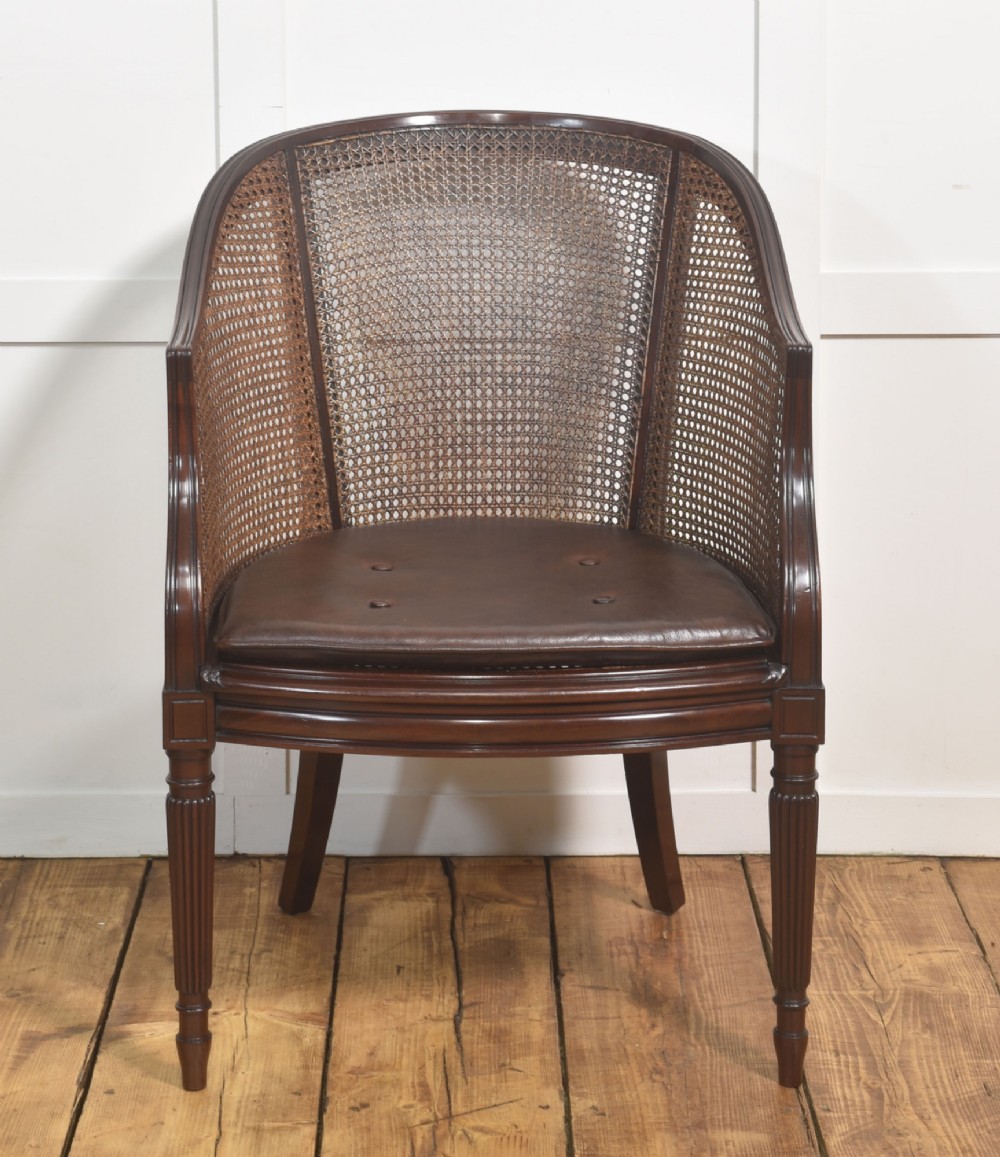 An Edwardian mahogany tub armchair featuring curved woven cane panels, a brown leather seat cushion, and fluted front legs. View V