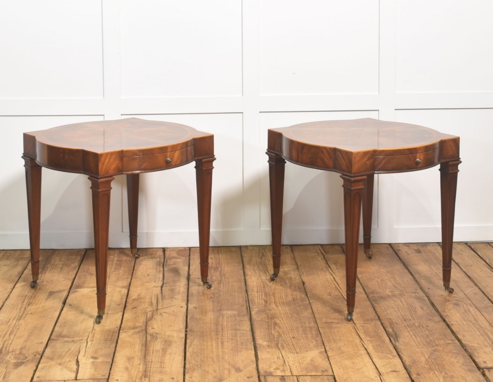 A pair of mahogany Sheraton Revival side tables featuring shaped inlaid tops, single frieze drawers, fluted tapered legs, and brass castors.