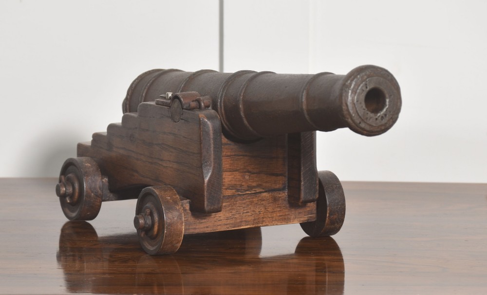 A miniature cast-iron signal cannon barrel mounted on a dark stepped oak wooden carriage with four wooden wheels.