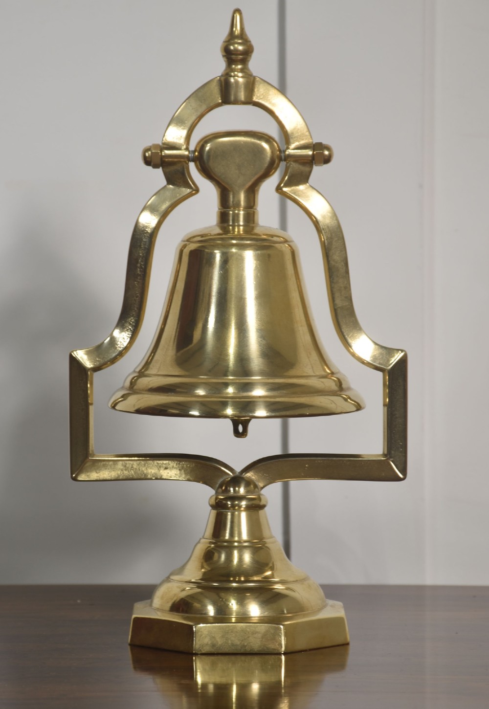 A polished brass table bell suspended within a shaped gantry frame on a tiered brass pedestal base.