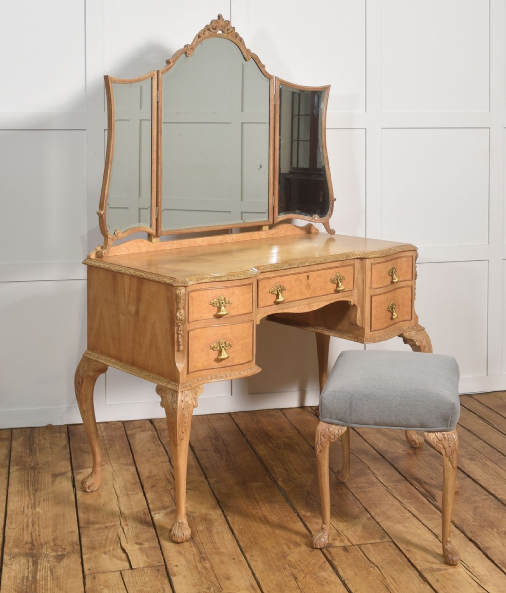 A light burr walnut dressing table featuring five drawers, brass handles, carved cabriole legs, a three-piece folding mirror, and a matching upholstered stool. View VI