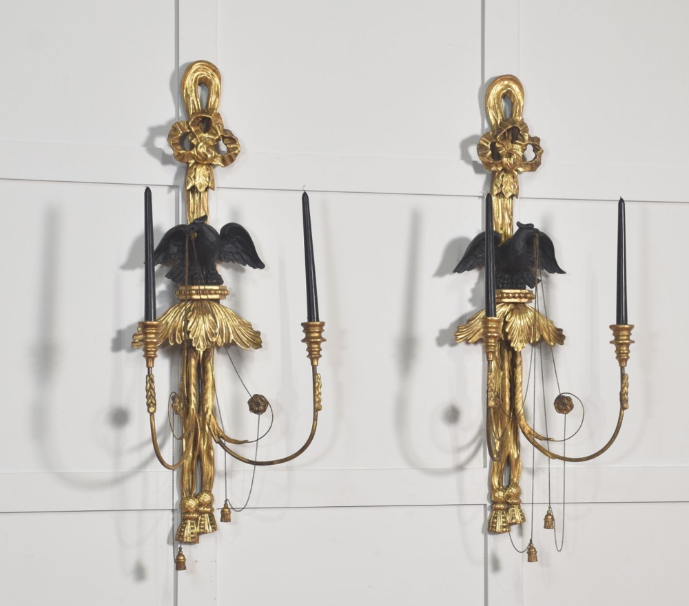 A pair of George III style giltwood wall sconces featuring carved ribbon crestings, dark eagle motifs, and twin candle arms. View X